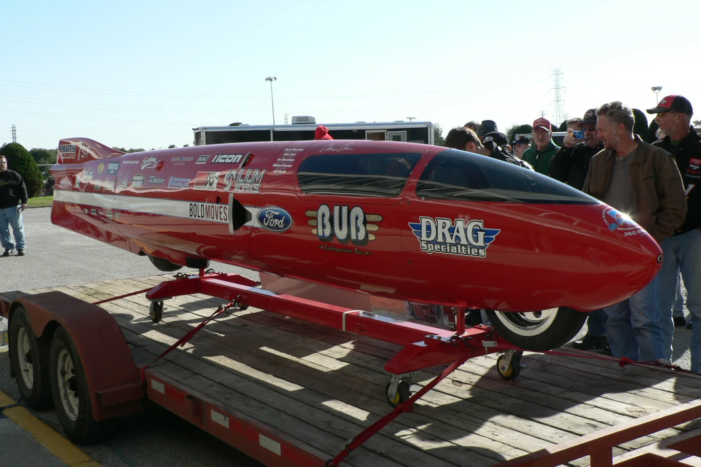 Czerwony pojazd wyścigowy o aerodynamicznym kształcie z licznymi logotypami sponsorów stoi na przyczepie, otoczony przez grupę ludzi obserwujących maszynę.