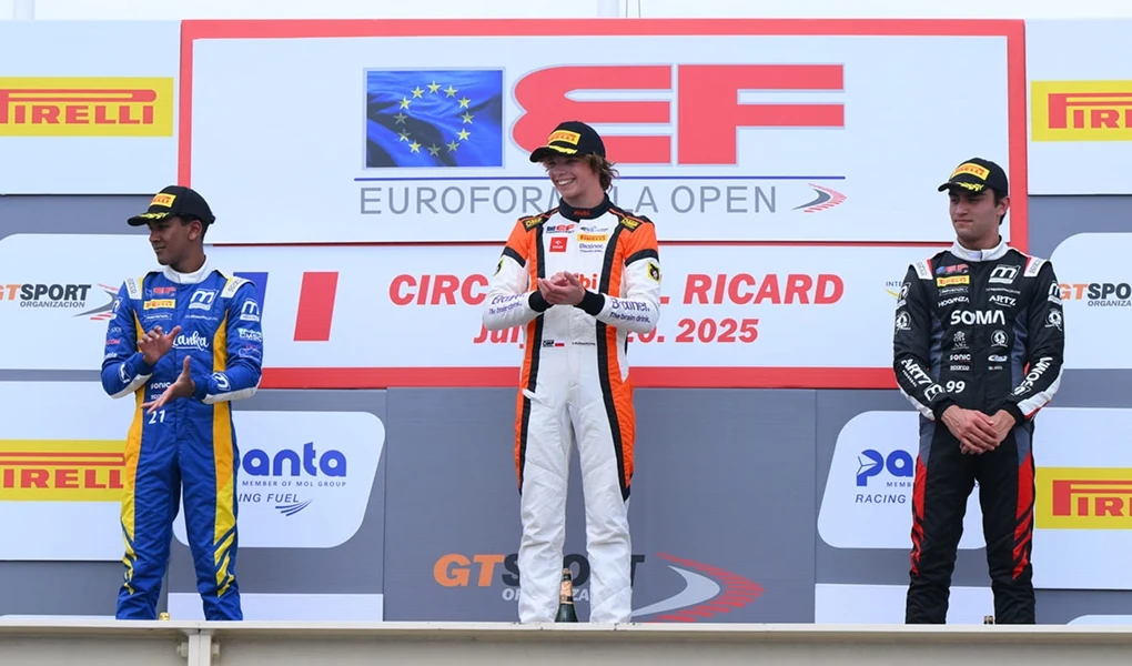 Laureaci wyścigu stoją na podium, środkowy zawodnik w biało-pomarańczowym kombinezonie trzyma trofeum, obok niego po obu stronach stoją pozostali kierowcy w niebieskim oraz czarnym stroju, tło z logiem i nazwą eliminacji Euroformula Open oraz sponsorami.