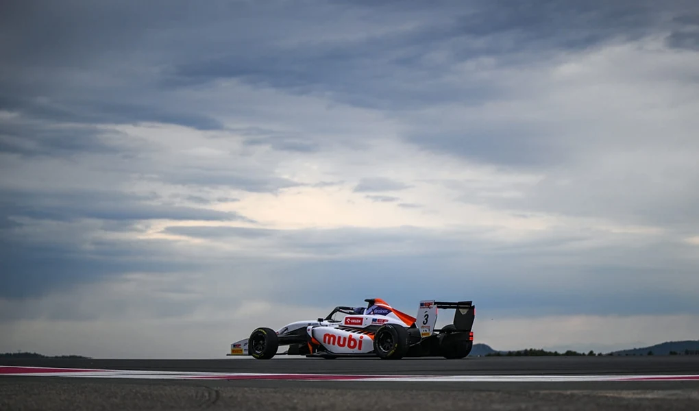 Samochód wyścigowy formuły jednoosobowej w dynamicznym ujęciu na torze przy pochmurnym niebie, z widocznym logo sponsora Mubi oraz numerem na skrzydle, w tle rozległy horyzont.