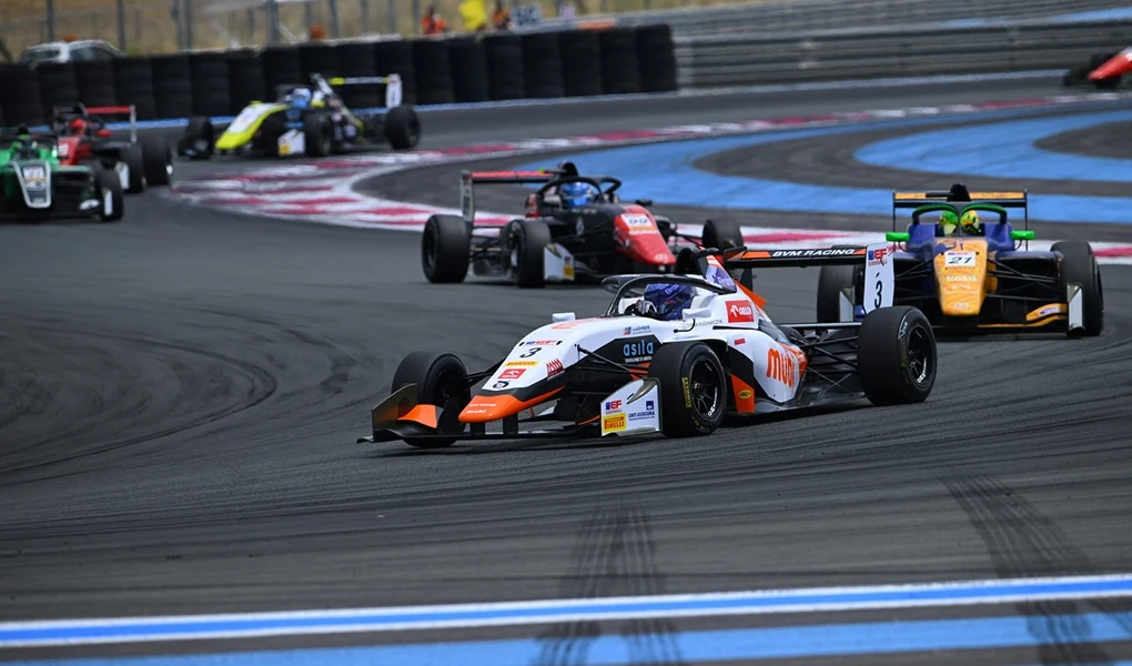 Kilka bolidów Formuły ściga się na asfalcie toru wyścigowego, prowadząc zaciętą rywalizację na zakręcie, otoczone barierami ochronnymi i pasami kolorowych oznaczeń toru.