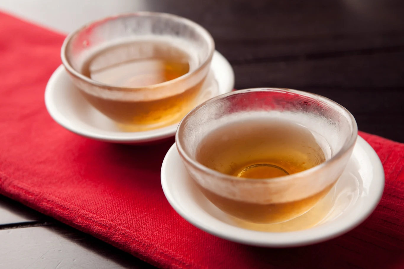Dlaczego warto pić ginseng tea Dwie przezroczyste filiżanki z herbatą ustawione na białych spodkach na czerwonym materiale, całość na ciemnym blacie, napój ma bursztynowy kolor.