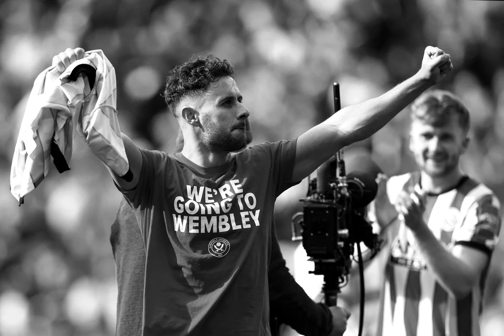 George Baldock Piłkarz z podniesioną ręką trzyma koszulkę, na jego własnej koszulce napis WE'RE GOING TO WEMBLEY, w tle widoczny drugi zawodnik bijący brawo i kamera.