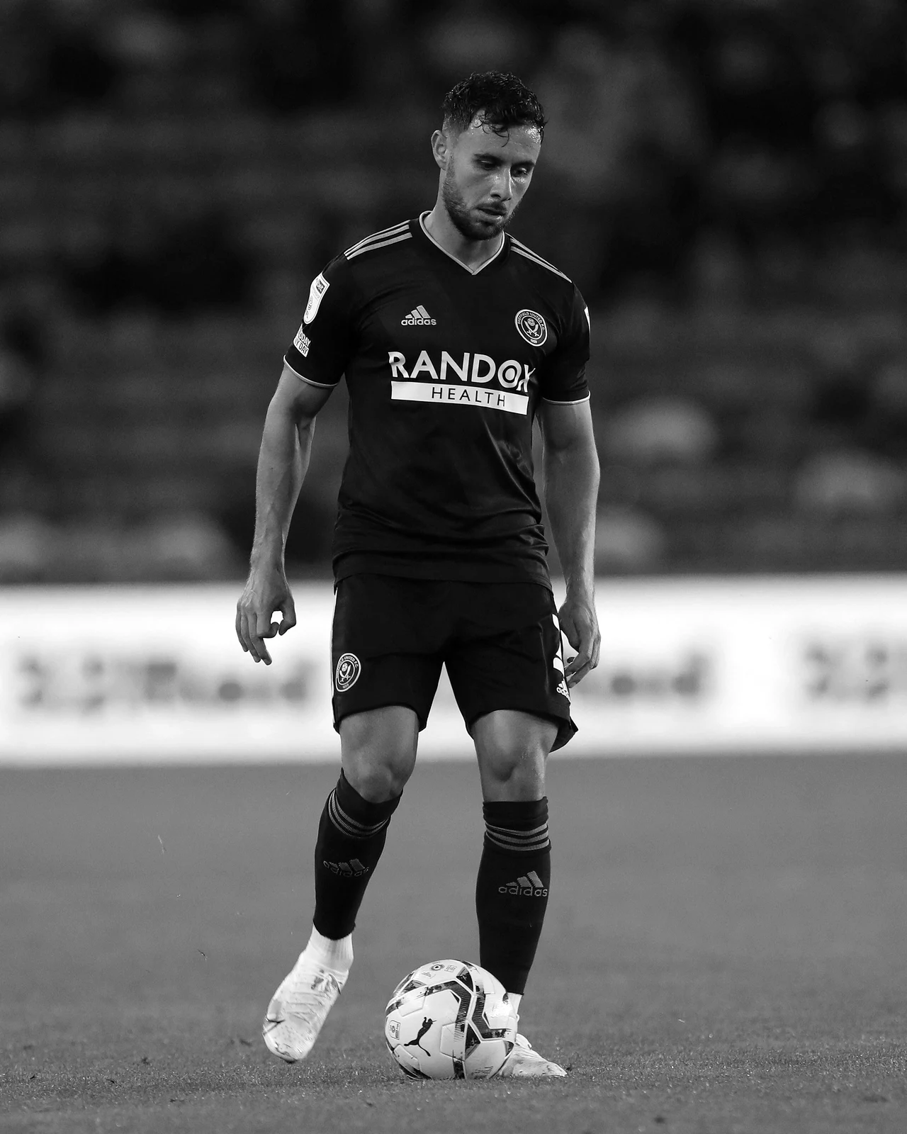 George Baldock Piłkarz w ciemnym stroju sportowym stoi na boisku, koncentrując się na piłce nożnej znajdującej się przed jego stopami; w tle rozmazane trybuny.