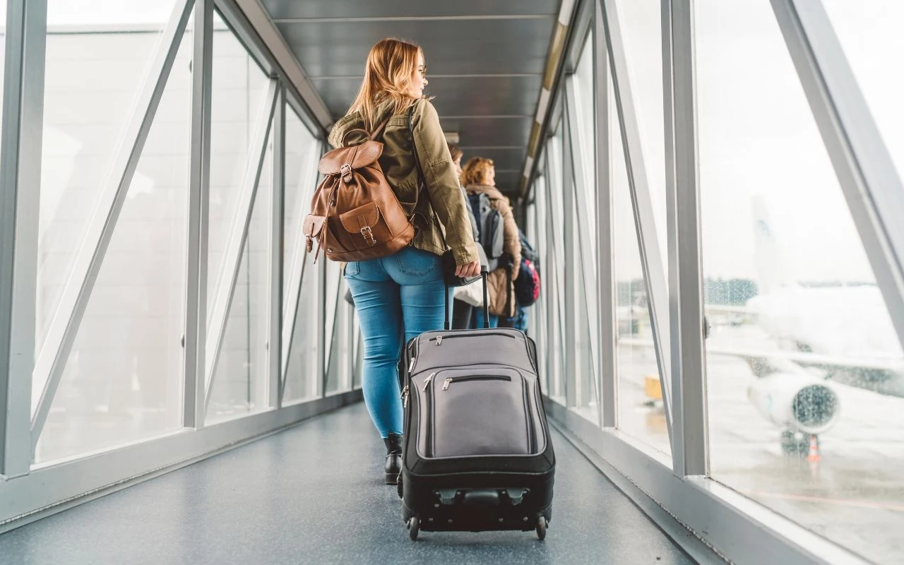 Kobieta z plecakiem i walizką na kółkach wchodzi do samolotu przez przeszklony rękaw lotniskowy, w tle widać samolot do którego się kieruje oraz inne osoby idące w tym samym kierunku.