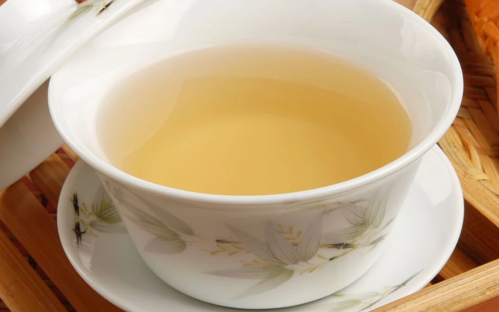 Porcelanowa filiżanka z jasnym naparem herbaty ustawiona na dopasowanym spodku ze wzorem kwiatowym, całość na tle drewnianej tacy.
