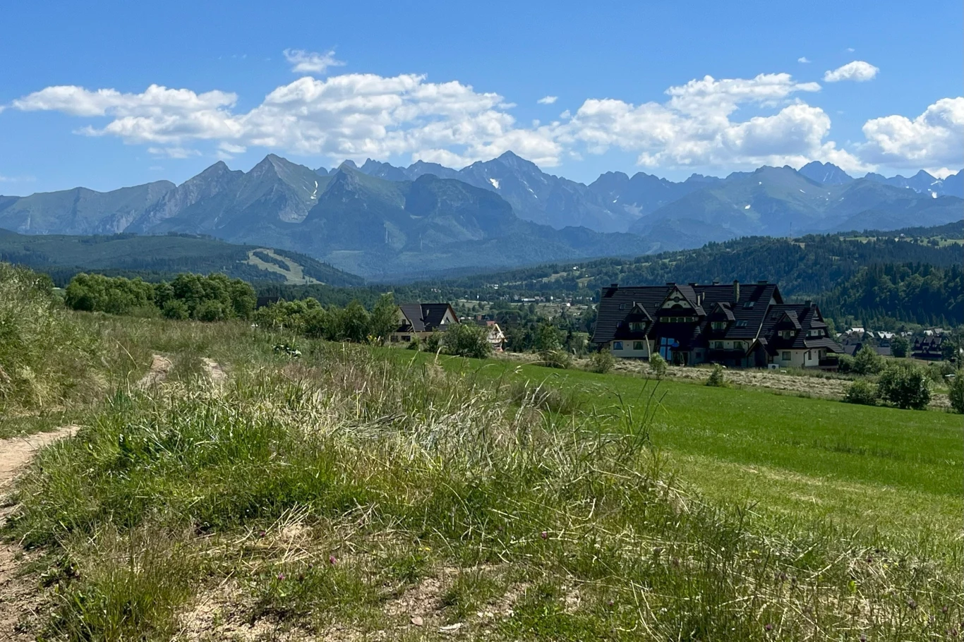 Górska panorama z zielonymi łąkami na pierwszym planie, domy w typowej dla regionu architekturze oraz w tle majestatyczne szczyty Tatr pod błękitnym niebem z chmurami.
