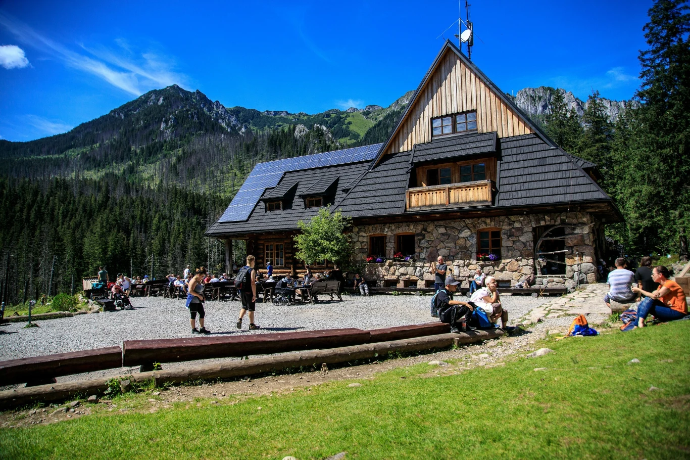 Schronisko na Hali Ornak. Oaza ciszy w Tatrach Zachodnich Górskie schronisko z kamienną elewacją i drewnianym dachem położone u podnóża lasu, otoczone górami, przed budynkiem turyści odpoczywają na ławkach i spacerują przy słonecznej pogodzie.