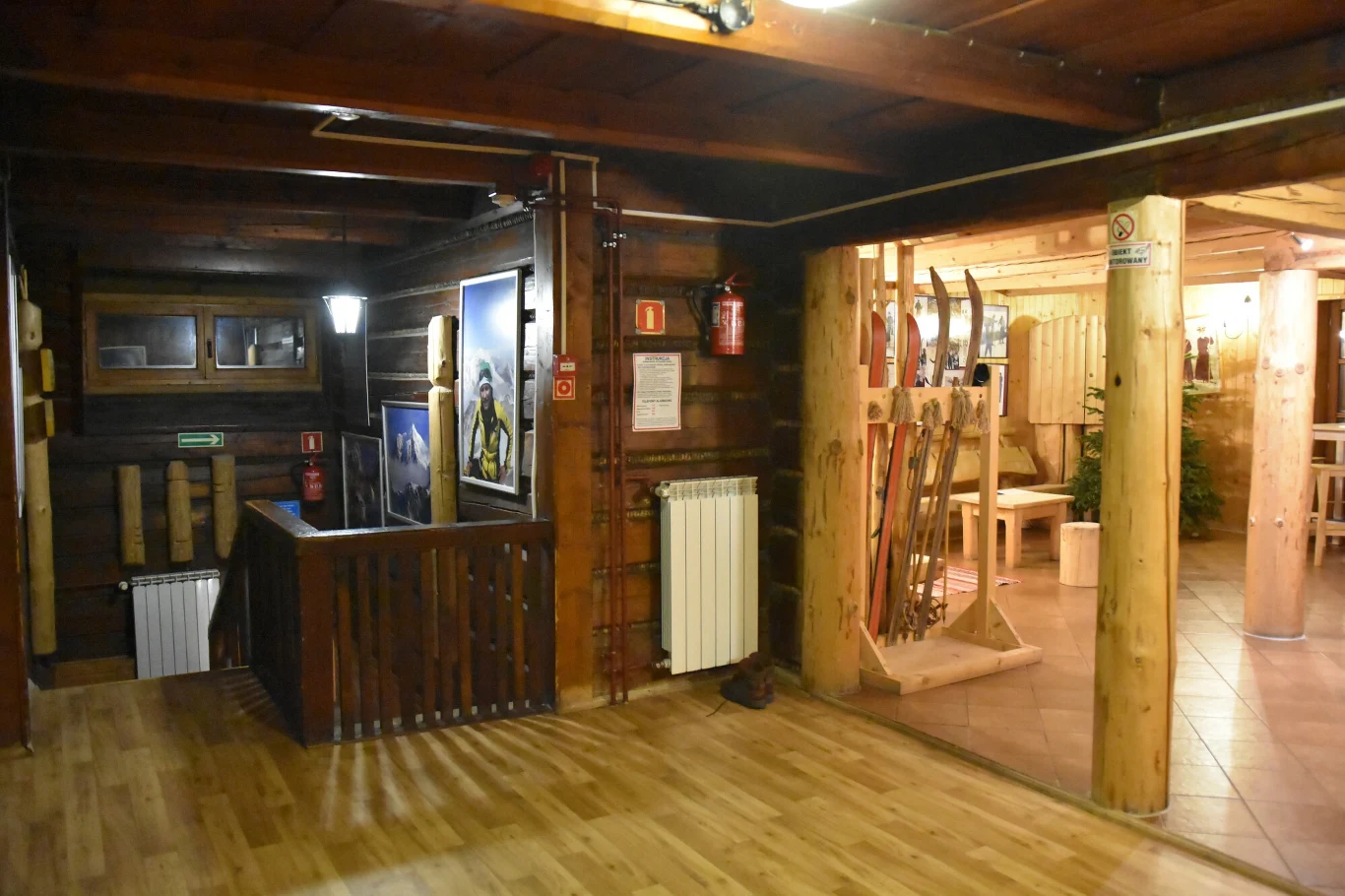 Wnętrze schroniska PTTK na Hali Ornak Przestrzeń wnętrza z drewnianymi ścianami i podłogą, w której znajduje się ekspozycja starych nart opartych o ścianę oraz elementy dekoracyjne nawiązujące do górskiej tematyki, oświetlona ciepłym światłem.