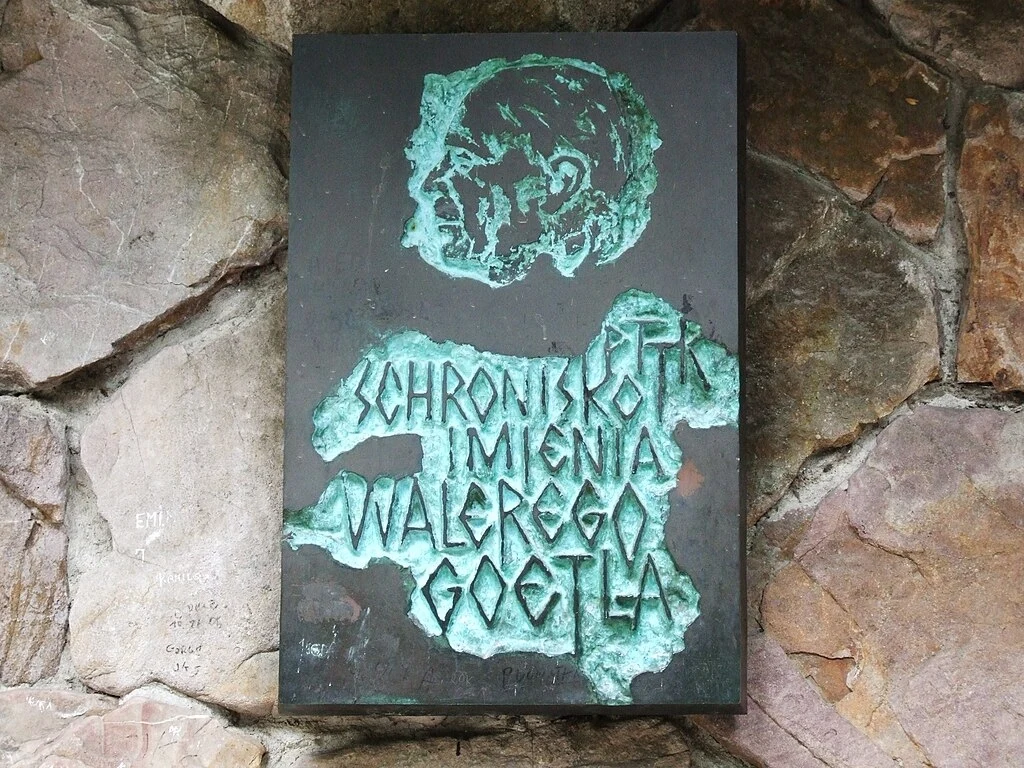 Tablica przy wejściu z nazwiskiem patrona Tablica pamiątkowa wykonana z metalu zamocowana na kamiennej ścianie, przedstawiająca wypukły profil mężczyzny oraz napisy informujące o schronisku PTTK imienia Walerego Goetla.