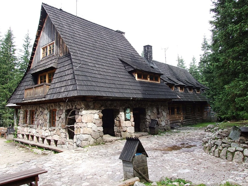 Widok od strony głównego wejścia, ze szlaku w Dolinie Kościeliskiej Duży góralski budynek z kamienia i drewna otoczony lasem świerkowym, masywny dach kryty gontem, w pobliżu kamienny krąg i drewniane ławki, spokojne otoczenie górskie.