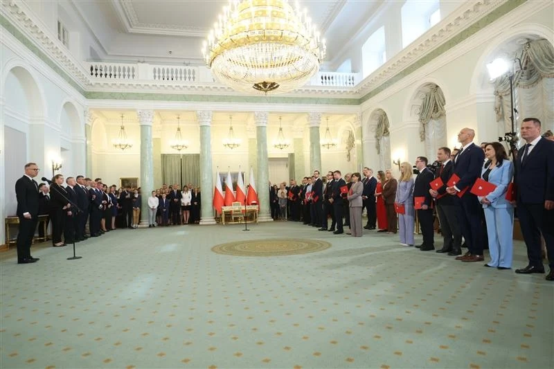 Zaprzysiężenie nowych ministrów w Pałacu Prezydenckim Duża sala z żyrandolem i kolumnami, w której zgromadziła się grupa osób ubranych formalnie, stojących po obu stronach pomieszczenia, naprzeciwko siebie; w głębi widoczne polskie flagi rozstawione za stołem.