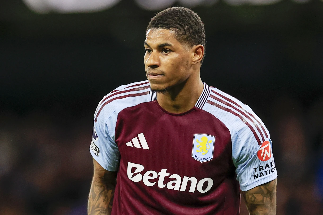 Marcus Rashford Piłkarz w bordowo-błękitnej koszulce klubu Aston Villa podczas meczu, skupiony i patrzący przed siebie, z widocznym logo sponsora i emblematem drużyny na koszulce.