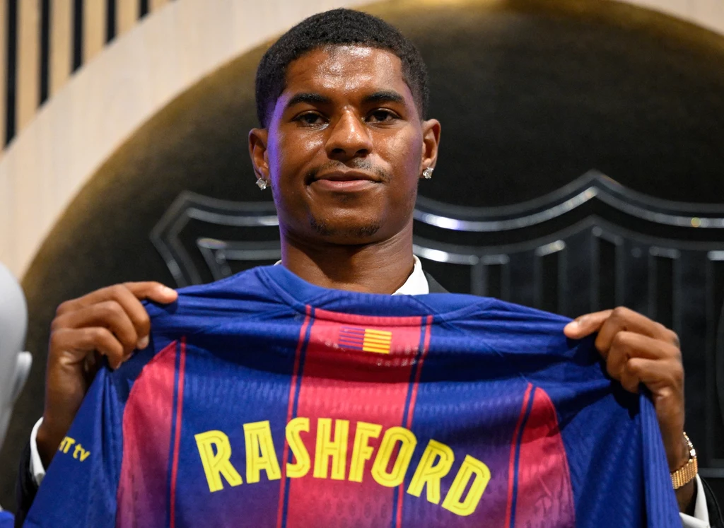 Marcus Rashford Mężczyzna prezentuje piłkarską koszulkę z nazwiskiem Rashford, stojąc na tle czarnego emblematu klubu sportowego.