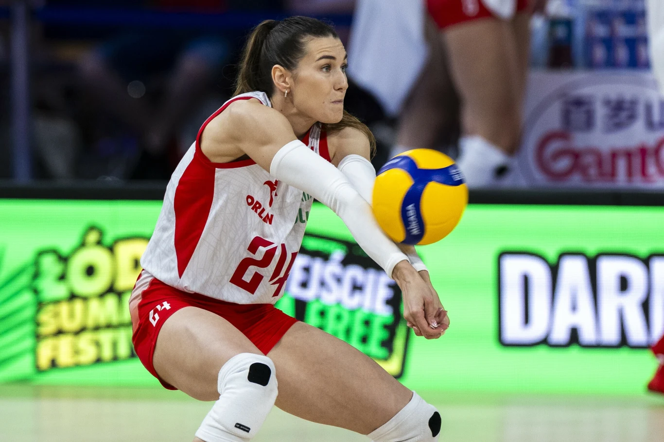 Zawodniczka w stroju siatkarskim wykonuje odbiór piłki podczas meczu, skupiona na precyzyjnym zagrywaniu, w tle widoczne elementy hali sportowej i reklamy.