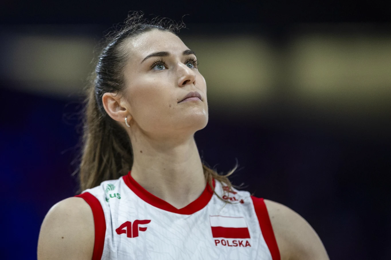 Kobieta w sportowym stroju reprezentacji Polski, patrząca w górę z wyrazem skupienia na twarzy, wyraźnie widoczny napis 'Polska' oraz logo sponsora na koszulce.