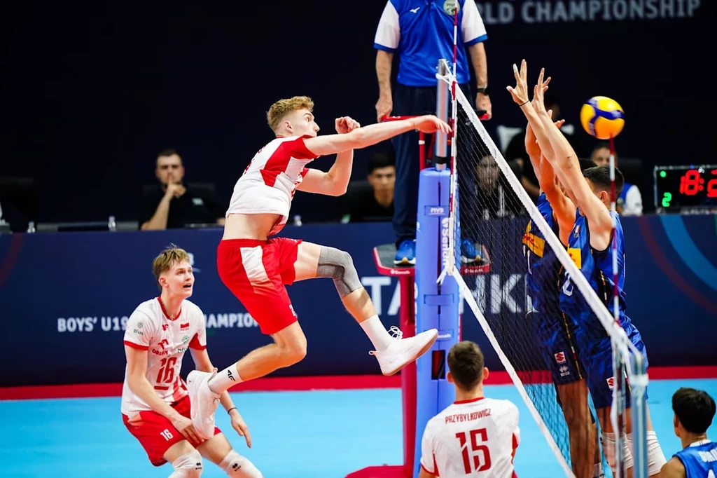 Mecz Polska - Włochy na MŚ U-19 Polski siatkarz wykonuje dynamiczny atak nad siatką podczas meczu, pozostali zawodnicy jednej i drugiej drużyny przygotowani do blokowania lub obrony, tło wypełnia logo mistrzostw świata i sędziowie.