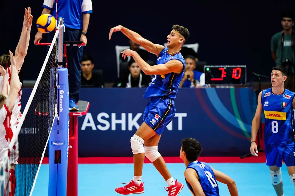 Kadr z meczu Polska - Włochy U19 Zawodnik drużyny grającej w niebieskich strojach na podskoku wykonuje atak siatkarski przy siatce, naprzeciw widoczni przeciwnicy starający się zablokować piłkę, tło stanowią kibice i sędziowie.