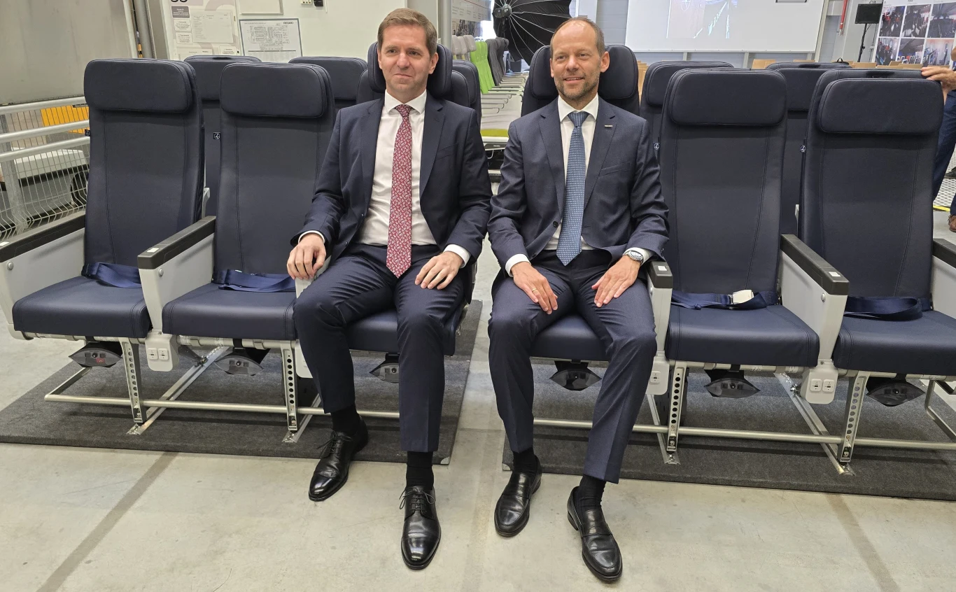 Michał Fijoł, prezes PLL LOT i Mark Hiller, szef Recaro, producenta foteli do samolotów polskiego przewoźnika. Dwóch mężczyzn w garniturach siedzi obok siebie na siedzeniach lotniczych, ustawionych w zakładzie przemysłowym lub hali produkcyjnej, co sugeruje testowanie lub prezentację elementów wnętrza samolotu.
