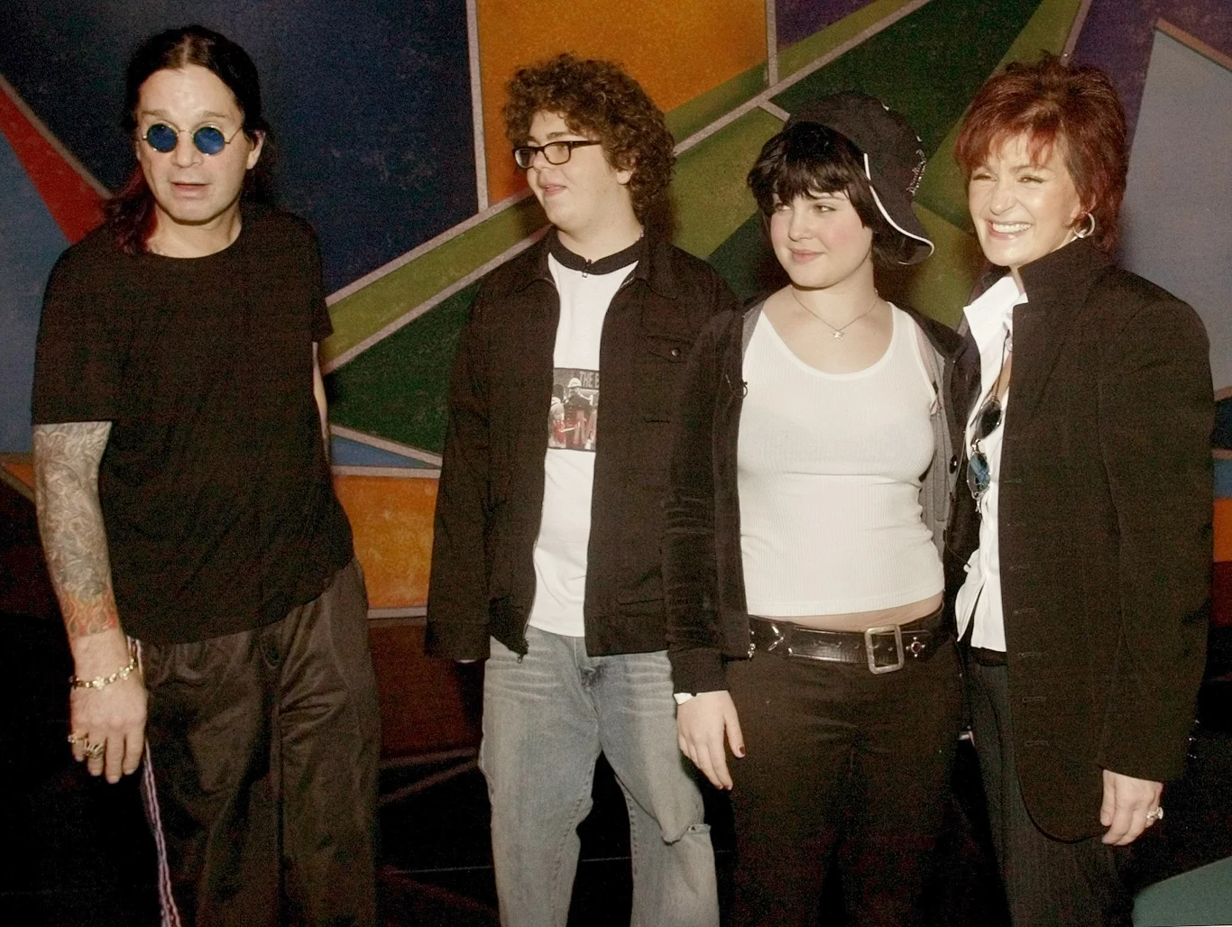 Rodzina Osbourne'ów w 2003 r. - od lewej: Ozzy, Jack, Kelly i Sharon Cztery osoby pozują razem na tle kolorowej, geometrycznej ściany; dwie z nich są ubrane na czarno, jedna nosi okulary przeciwsłoneczne, druga czapkę z daszkiem, pozostałe dwie uśmiechają się.