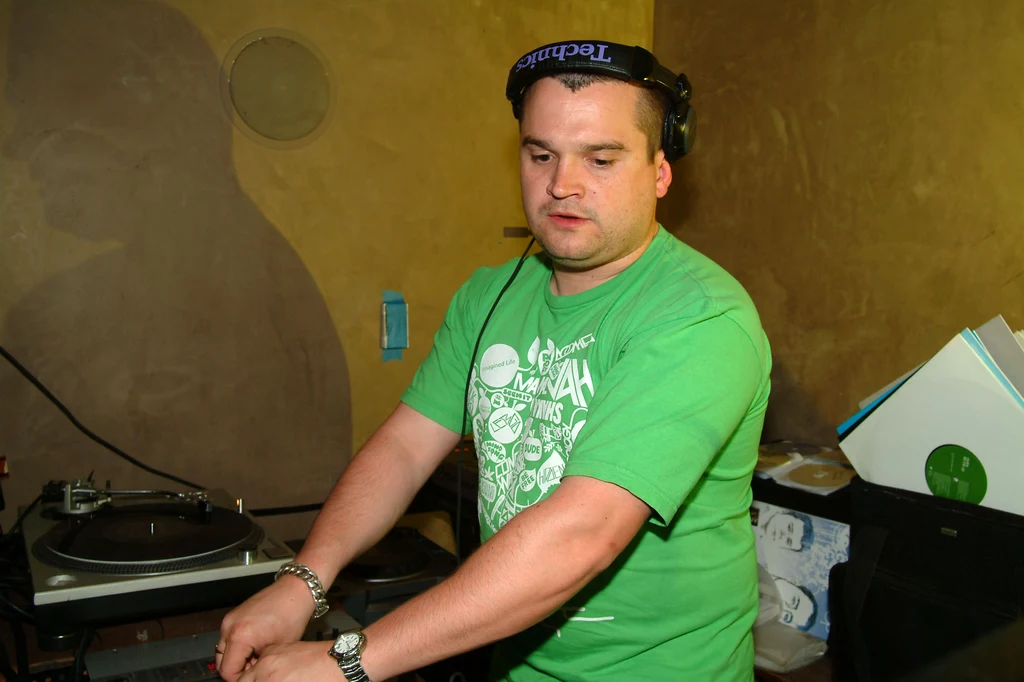 Louis Osbourne w roli DJ-a w 2005 r. DJ w zielonej koszulce miksujący muzykę przy konsoli DJ-skiej z gramofonem, słuchawkami na głowie oraz sprzętem muzycznym w tle.