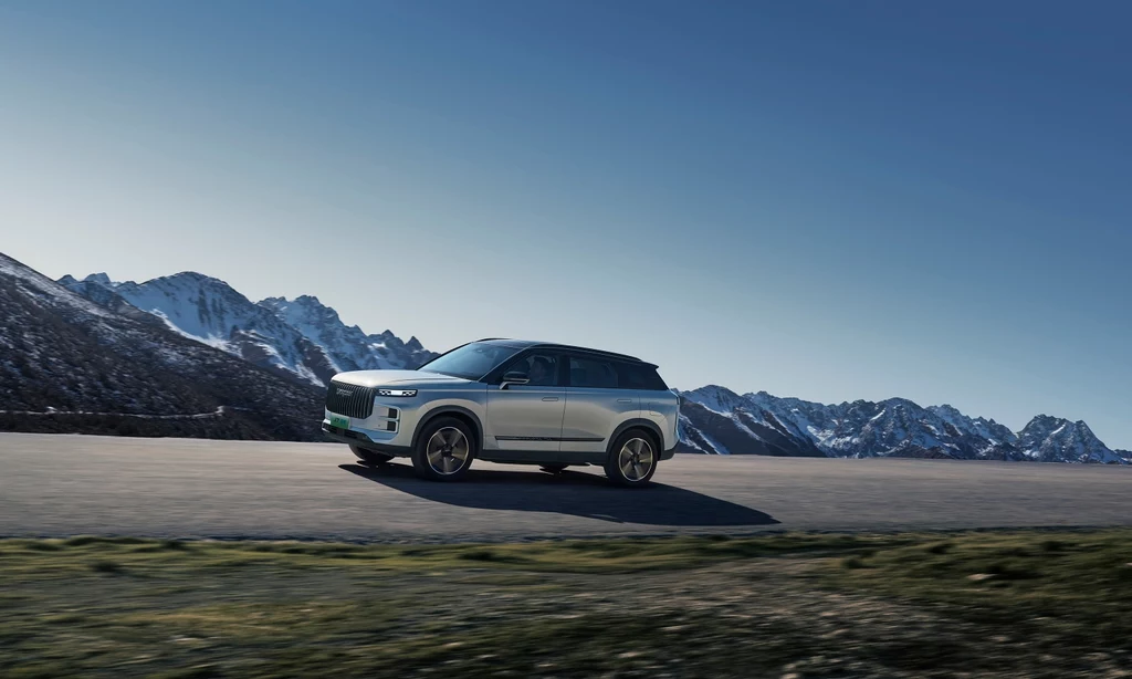 Jaecoo 7 Super Hybrid objęty jest 7-letnią gwarancją lub do 150 000 km Srebrny SUV jedzie pustą, asfaltową drogą, położoną u podnóża ośnieżonych gór przy jasnym, bezchmurnym niebie.