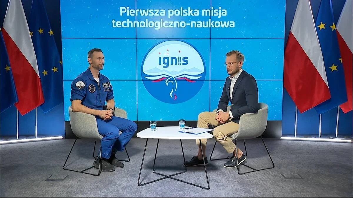 Sławosz Uznański-Wiśniewski. Pierwszy wywiad po powrocie do Polski. Dwóch mężczyzn podczas wywiadu telewizyjnego, jeden z nich w niebieskim kombinezonie astronauty, drugi w garniturze, na tle ekranu z logotypem i napisem o pierwszej polskiej misji technologiczno-naukowej, po bokach ustawione polskie i unijne flagi.