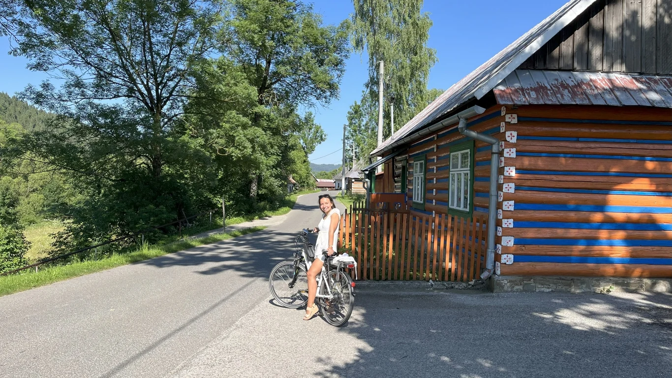 Kobieta w letniej sukience stoi przy rowerze na wiejskiej drodze obok drewnianego, pomalowanego w niebieskie pasy domu, otoczona zielonymi drzewami i jasnym, słonecznym niebem.