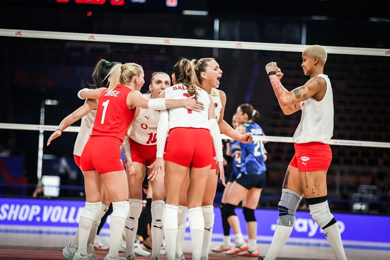 Grupa siatkarek w czerwono-białych strojach celebruje punkt na boisku, wyrażając radość przez okrzyki i gesty, jedna z zawodniczek unosi zaciśniętą pięść, a w tle widać siatkarkę drużyny przeciwnej w niebieskim stroju.