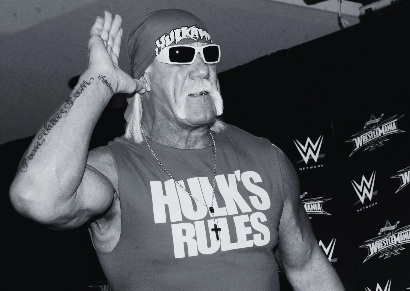 Mężczyzna w ciemnych okularach, bandanie i koszulce bez rękawów z napisem HULK'S RULES wykonuje gest dłonią przy uchu, w tle widoczne logo WWE oraz plansza z napisem WrestleMania.