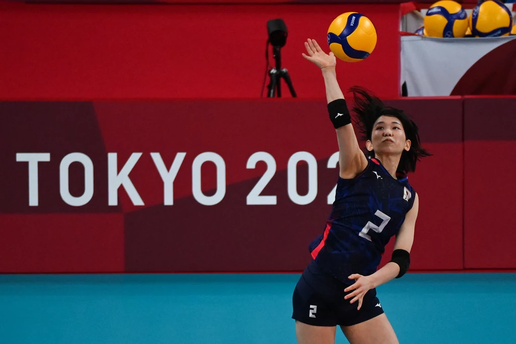 Zawodniczka siatkarska w ciemnogranatowym stroju z numerem 2 wykonuje serw na hali podczas igrzysk olimpijskich w Tokio, w tle fragment napisu TOKYO 2020 i piłki do siatkówki.