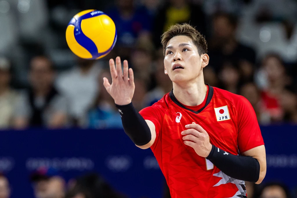 Japoński siatkarz w czerwonym stroju z numerem i flagą Japonii przygotowuje się do zagrania piłką podczas meczu, w tle rozmazana publiczność.