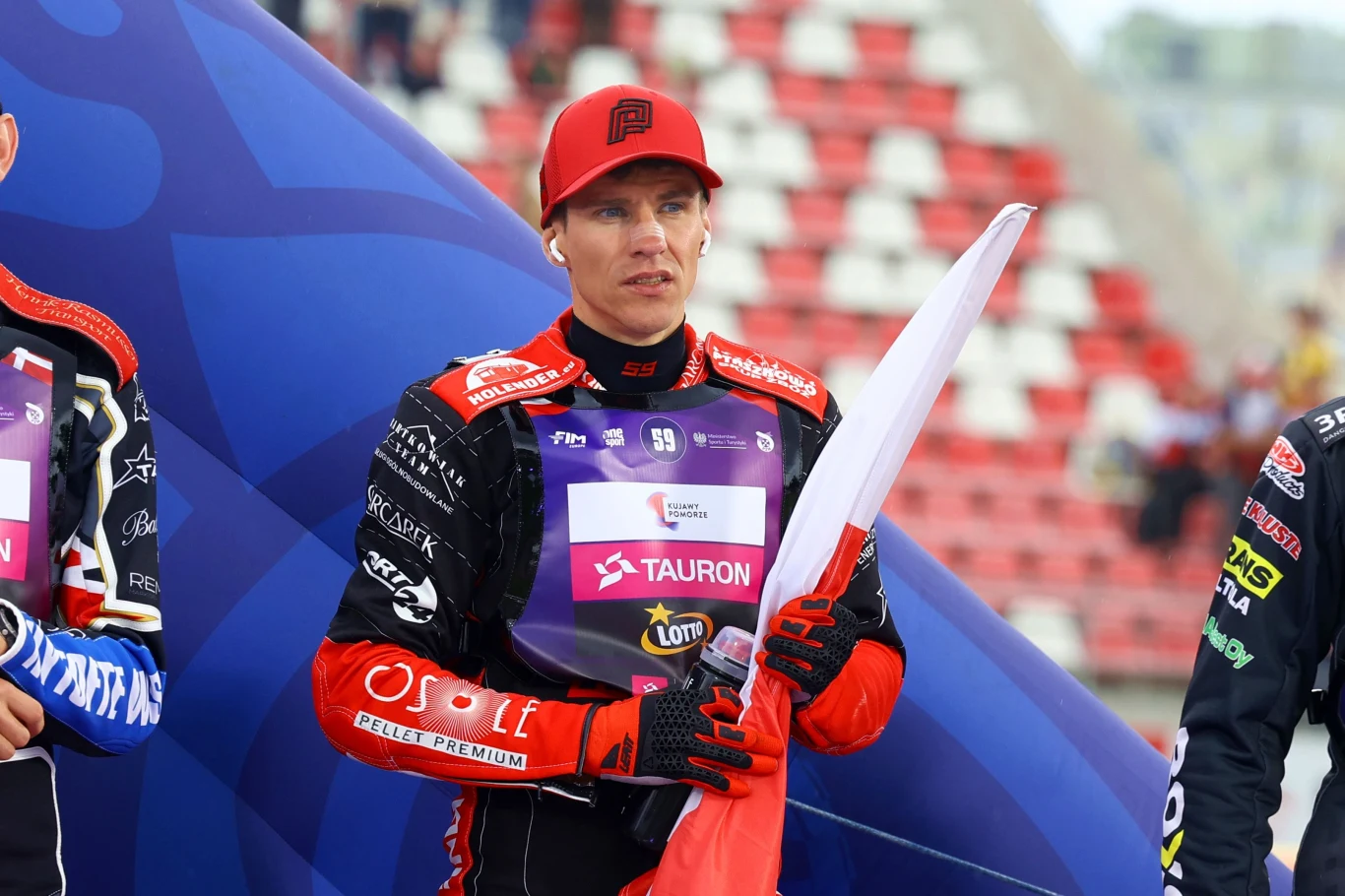 Przemysław Pawlicki Zawodnik żużlowy w stroju sportowym i czerwonej czapce trzyma biało-czerwoną flagę, stojąc na stadionie z trybunami w tle, obok innych sportowców.