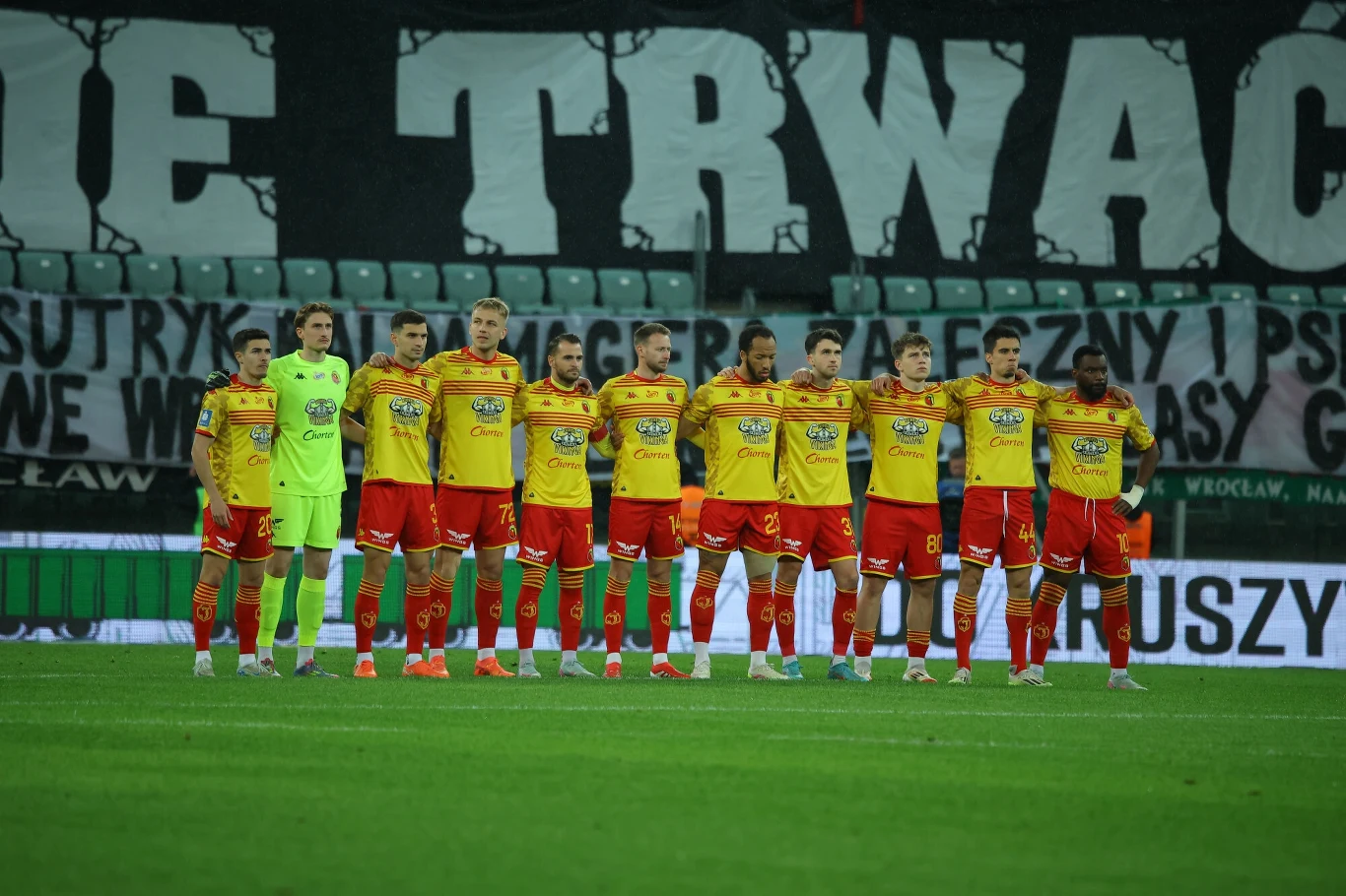 Piłkarze drużyny w żółto-czerwonych strojach stojący ramię w ramię na zielonej murawie stadionu podczas przygotowań do meczu, w tle widoczny transparent oraz puste krzesełka trybun.