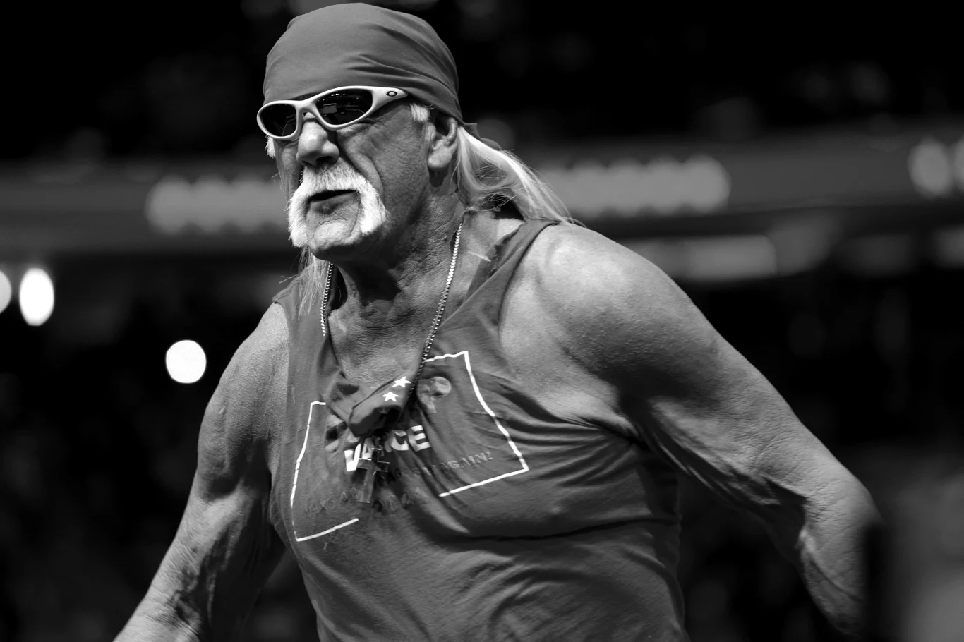 Hulk Hogan Starszy mężczyzna w bandanie i okularach przeciwsłonecznych w sportowej, muskularnej sylwetce, ubrany w koszulkę bez rękawów, sfotografowany w dynamicznej pozie na tle rozmytego tłumu.