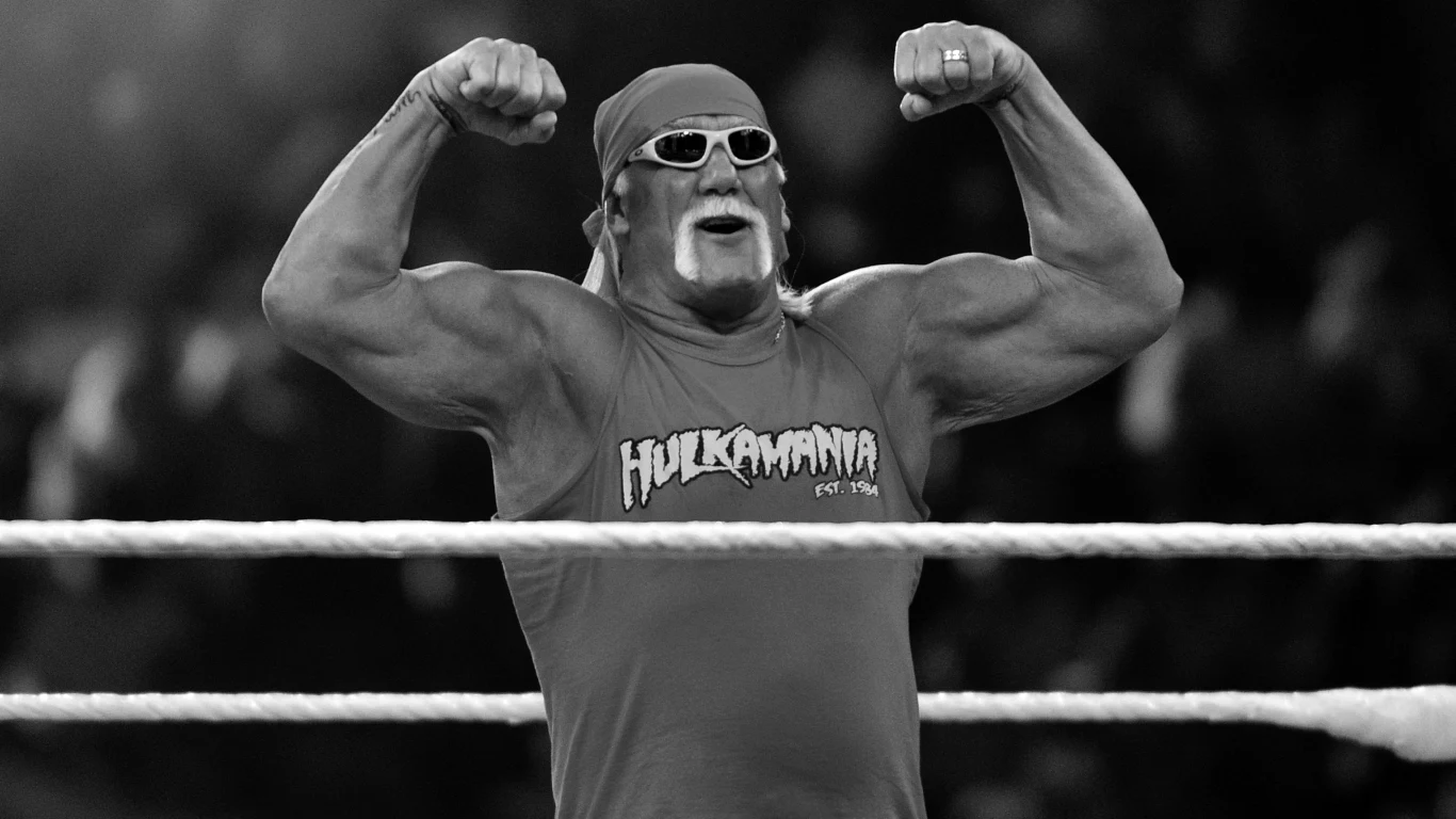 Muskularny mężczyzna z opaską na głowie i charakterystycznym wąsem pozuje, prężąc bicepsy na ringu, ubrany w koszulkę z napisem 'Hulkamania' i okulary przeciwsłoneczne.