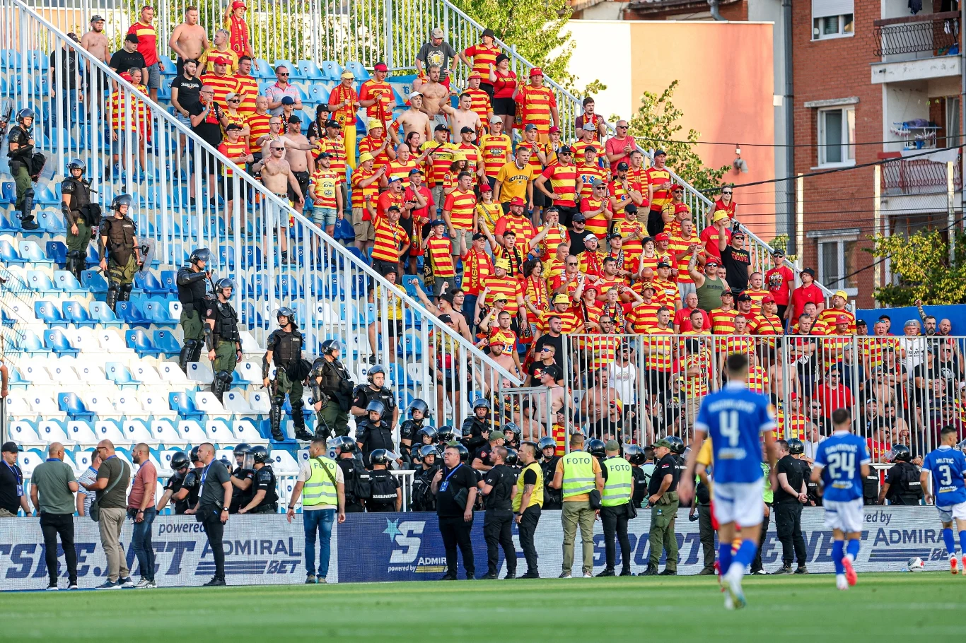 Trybuny stadionu wypełnione kibicami ubranymi w żółto-czerwone koszulki, część z nich stoi, część siedzi, widoczni także policjanci i ochrona stojący wzdłuż sektorów, na boisku piłkarze w niebieskich strojach.
