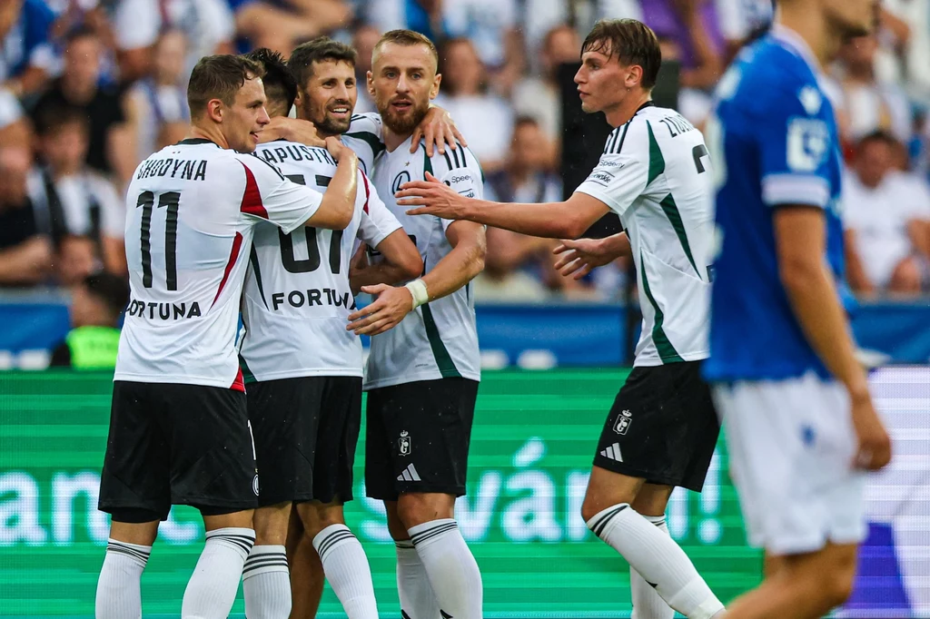 Gratulacje odbiera Bartosz Kapustka. W tym momencie było 1:1 Kilku piłkarzy w białych strojach drużyny Legii Warszawa świętuje zdobycie gola, jeden z zawodników obejmuje kolegę z zespołu, w tle znajduje się boisko oraz zawodnicy drużyny przeciwnej ubrani na niebiesko.