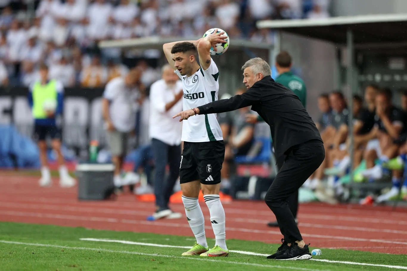 Zawodnik w stroju sportowym przygotowuje się do wykonania wrzutu z autu podczas meczu piłki nożnej, obok trener udzielający wskazówek; w tle ławka rezerwowych oraz kibice na trybunach.