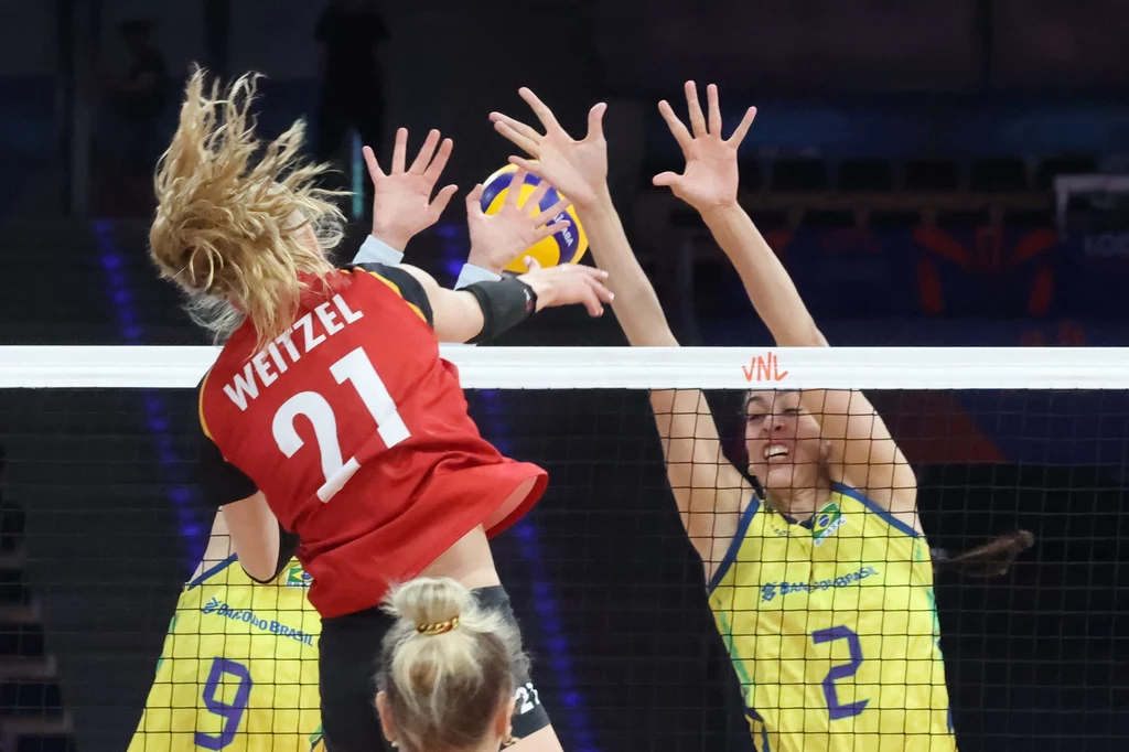 Brazylia - Niemcy Zawodniczka w czerwonej koszulce z numerem 21 atakuje piłkę przy siatce, naprzeciwko dwie siatkarki w żółtych strojach próbują zablokować atak, widoczne dynamiczne emocje sportowe i współzawodnictwo.