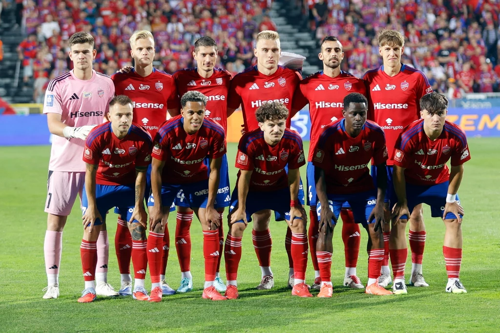 Jedenastu piłkarzy w czerwonych i niebieskich strojach drużyny sportowej ustawia się do wspólnego zdjęcia drużynowego na stadionie przed rozpoczęciem meczu, a w tle widownia z kibicami.