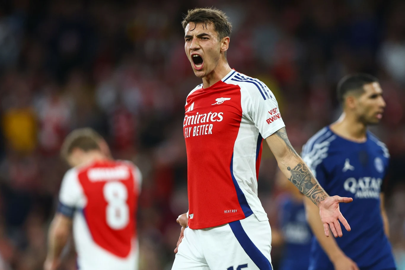 Jakub Kiwior Piłkarz drużyny Arsenal w czerwono-białej koszulce gestykuluje i krzyczy, pokazując silne emocje podczas meczu, w tle widoczni są inni zawodnicy oraz rozmyte trybuny.