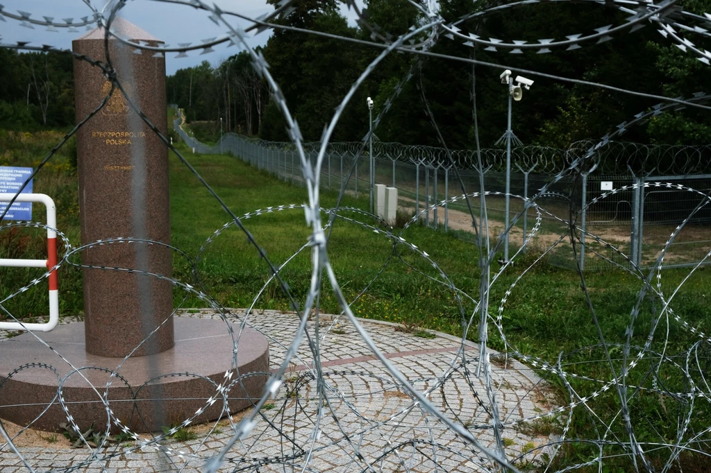 Płot z drutu kolczastego i zasieków oddzielający teren, w tle pomnik graniczny oraz kamery monitoringu, całość otoczona zielenią i lasem.
