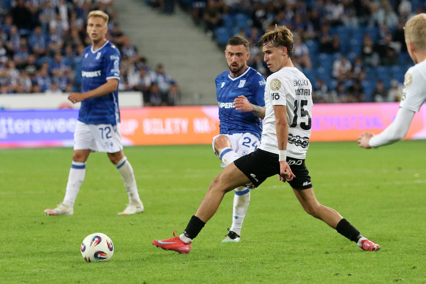 Lech Poznań pobił własny rekord strzelecki Dwóch piłkarzy walczy o piłkę podczas meczu piłki nożnej na stadionie, jeden zawodnik zakłada wślizg próbując przejąć piłkę od przeciwnika z niebieskiej drużyny, w tle widać innych zawodników oraz trybuny pełne kibiców.