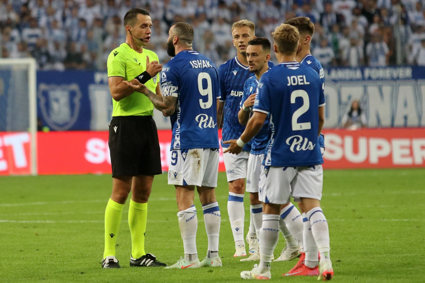 Kontrowersji w meczu Lech Poznań - Breidablik nie brakowało Grupa piłkarzy w niebieskich strojach podchodzi do sędziego ubranego w żółtą koszulkę i czarne spodenki na tle kibiców na stadionie, trwa wymiana opinii dotycząca przebiegu meczu.