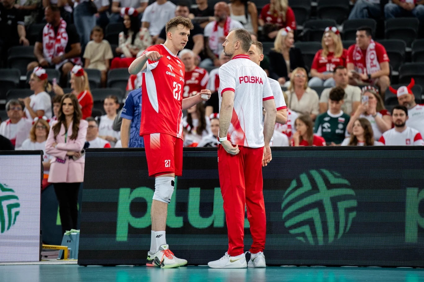 Dwóch mężczyzn w sportowych strojach reprezentacji Polski rozmawia na boisku siatkarskim, wokół nich zgromadzona publiczność, w tle widoczne banery sponsora oraz kibice w biało-czerwonych barwach.
