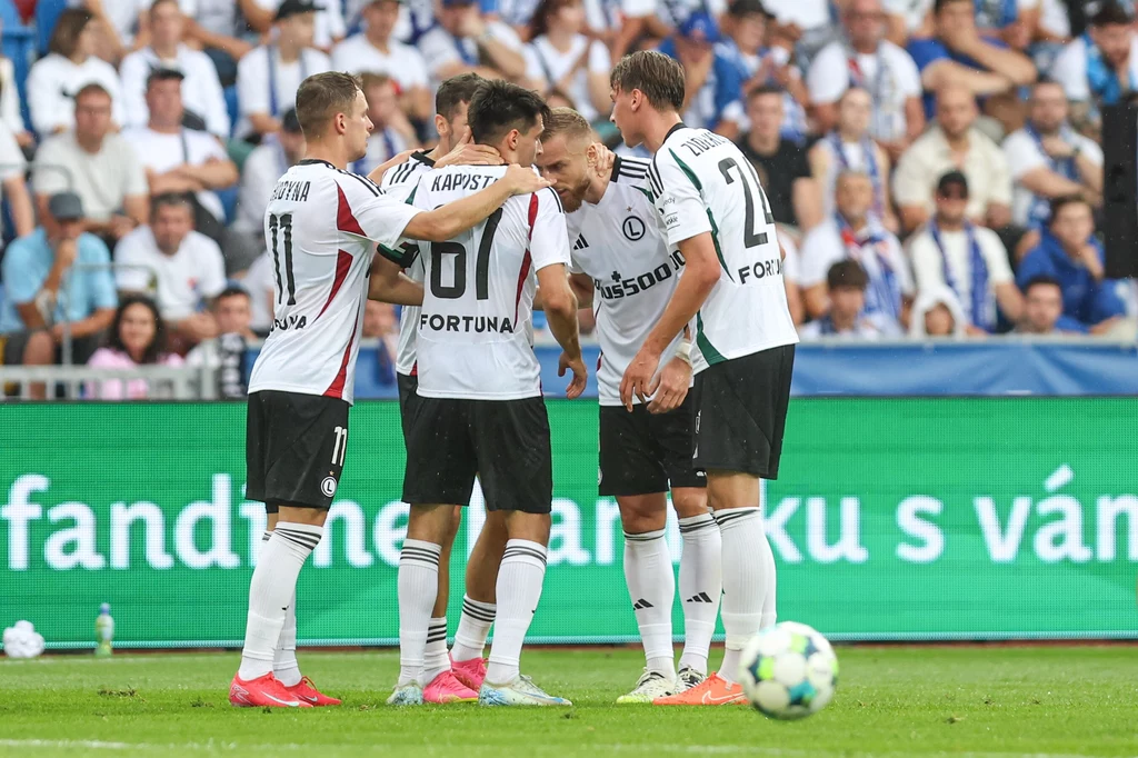 Legia Warszawa Grupa piłkarzy w białych koszulkach z czarnymi i czerwonymi detalami stoi razem na boisku podczas meczu, omawiając taktykę na tle kibiców i elektronicznej tablicy reklamowej.