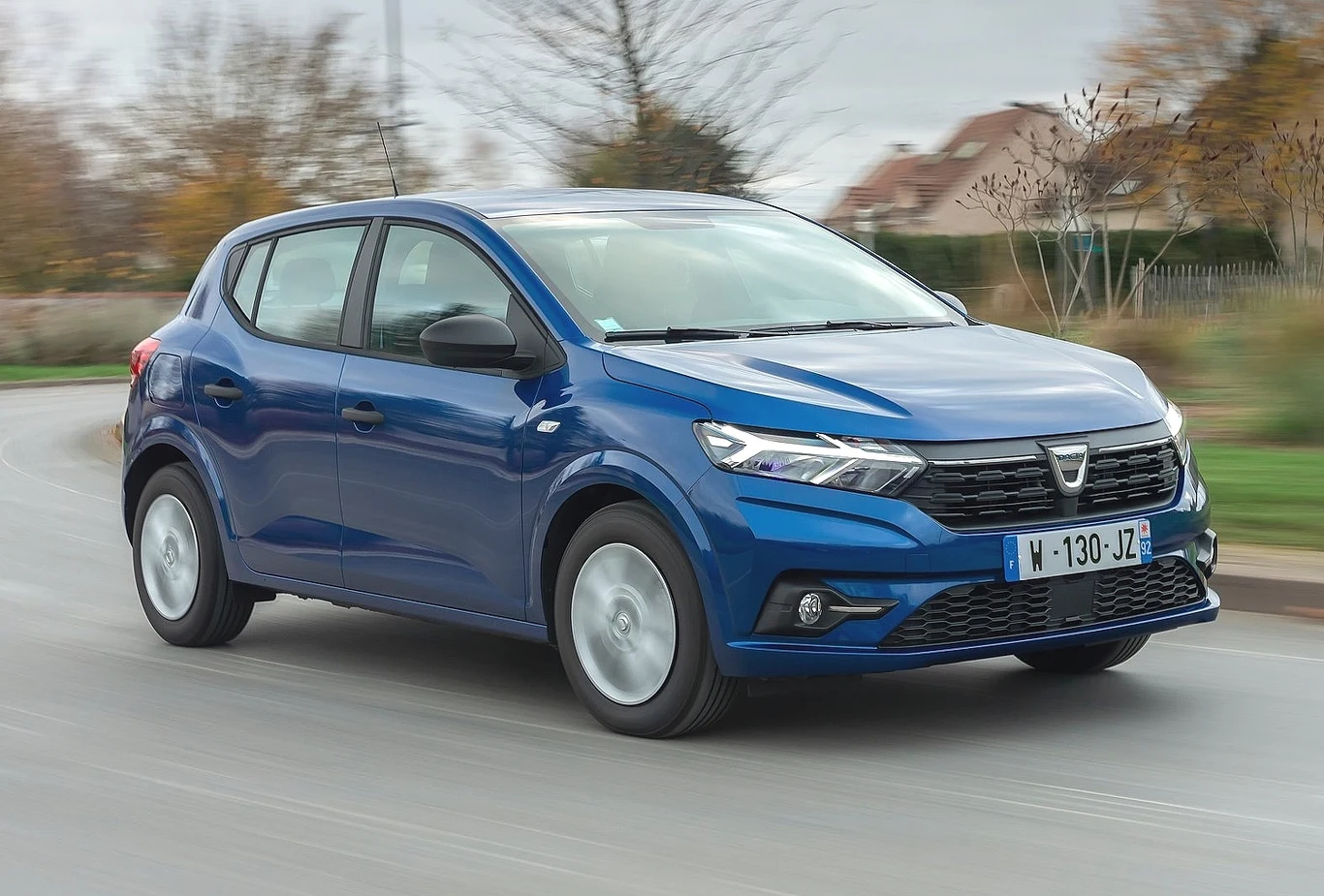 Niebieski samochód Dacia Sandero najnowszej generacji porusza się ulicą w otoczeniu jesiennych drzew oraz rozmytych domów w tle.