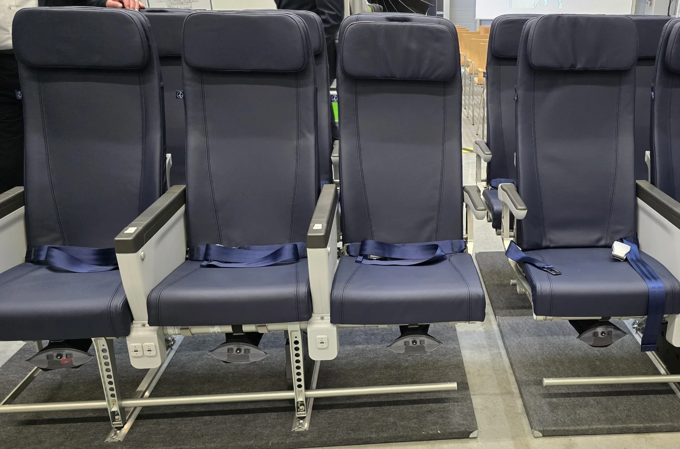 Fotele Recaro trafią do 13 Boeingów 737 MAX 8 polskiego LOT-u, a także do nowych Airbusów A220. Rząd ciemnych, nowoczesnych foteli lotniczych z pasami bezpieczeństwa w otoczeniu przemysłowego wnętrza, siedzenia ustawione na metalowych listwach, widoczna część podłogi i elementów montażowych.