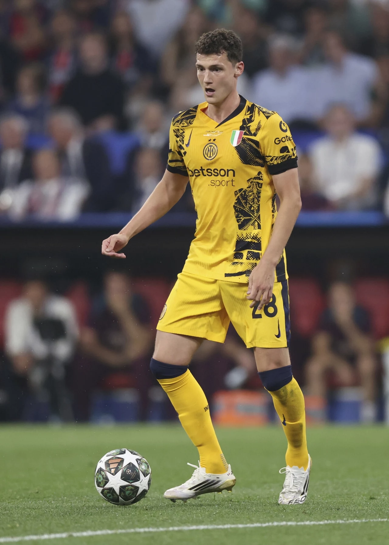 Benjamin Pavard Piłkarz w żółto-czarnym stroju drużynowym stoi na boisku podczas meczu, z piłką u nóg, skupiony na grze, w tle rozmazana publiczność stadionowa.