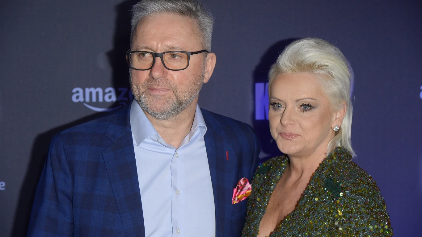 Jerzy Brzęczek z żoną Marzeną Para w eleganckich strojach wieczorowych pozuje wspólnie na tle ciemnego baneru z logotypami.